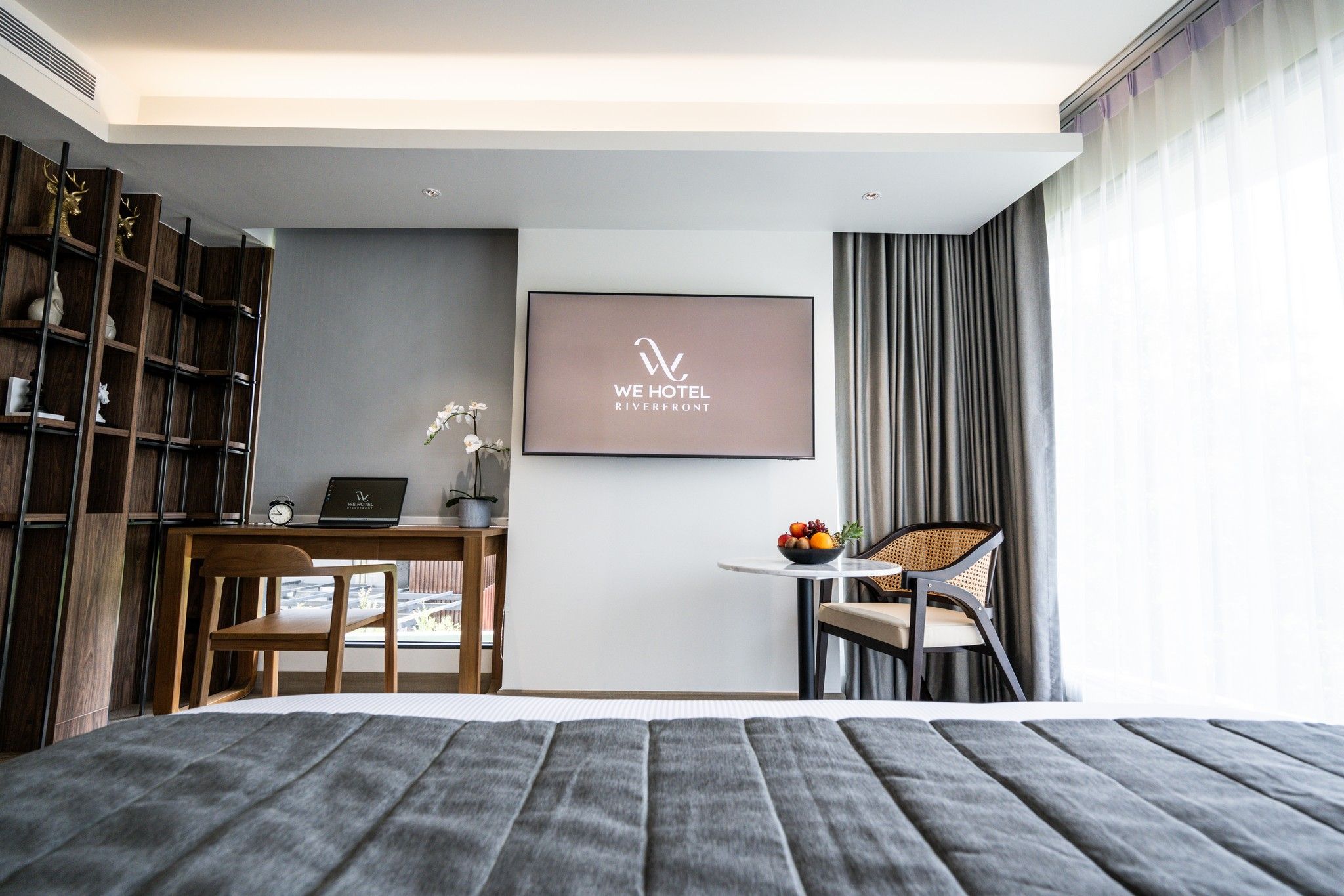 Habitación de hotel moderna con cama, escritorio con computadora portátil, pequeña mesa con fruta, una silla y estantería.
