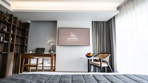 Ein modernes Hotelzimmer mit Bett, Schreibtisch mit Laptop, einem kleinen Tisch mit Obst, einem Sessel und einem Bücherregal.