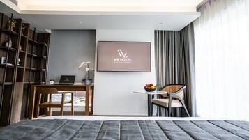 Habitación de hotel moderna con cama, escritorio con computadora portátil, pequeña mesa con fruta, una silla y estantería.