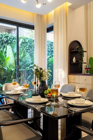 A dining area with a table set for four, surrounded by chairs, with a view of lush greenery through large windows. - Staymaster Villa Novara - 3BHK  (Vagator)