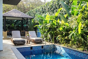 A pool area with two lounge chairs, a covered seating area, and lush tropical plants. - Staymaster Villa Novara - 3BHK  (Vagator)