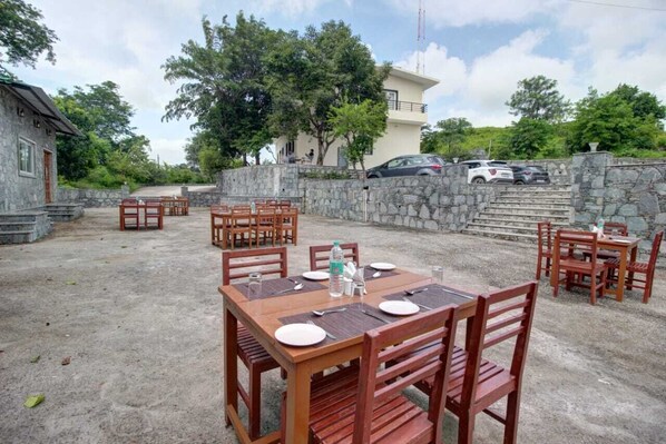 An outdoor dining area with stone walls, wooden tables and chairs, and a view of a building and parked cars. - Takhat villa kumbhalgarh (Kumbhalgarh)