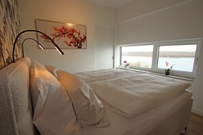A bedroom with a bed, a lamp, a window with a view, and a painting on the wall.