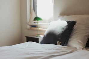 Ein ordentlich bezogenes Bett mit weißen Leinentüchern, ein schwarzes Kopfkissen mit Etikett und eine grüne Lampe auf einem Nachttisch.