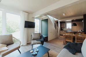 A modern living room with a sofa, chairs, a coffee table, and a TV. There is a staircase leading to a kitchen area with wooden cabinets and a dining table. - stilvolles skandinavisches XXL Familienferienhaus (Sagard)