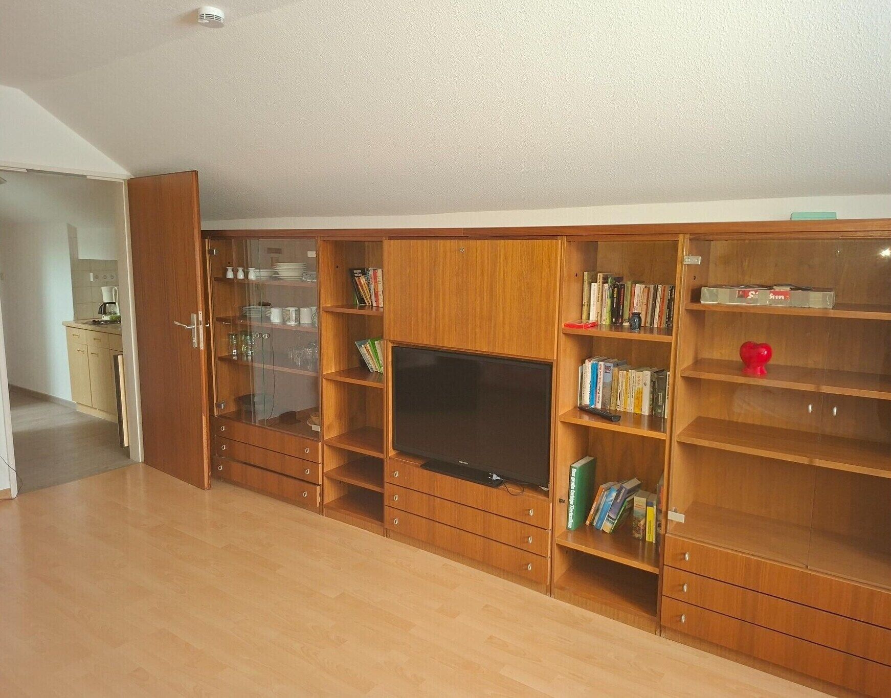 A spacious living room with a large wooden entertainment unit, a flat-screen TV, and a kitchen area visible through an open door.