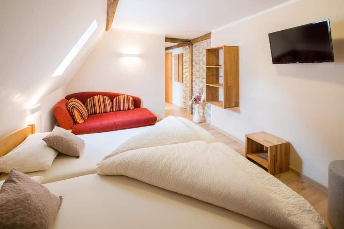 A bedroom with a red sofa, a bed with white pillows, a TV mounted on the wall, and a wooden shelf.