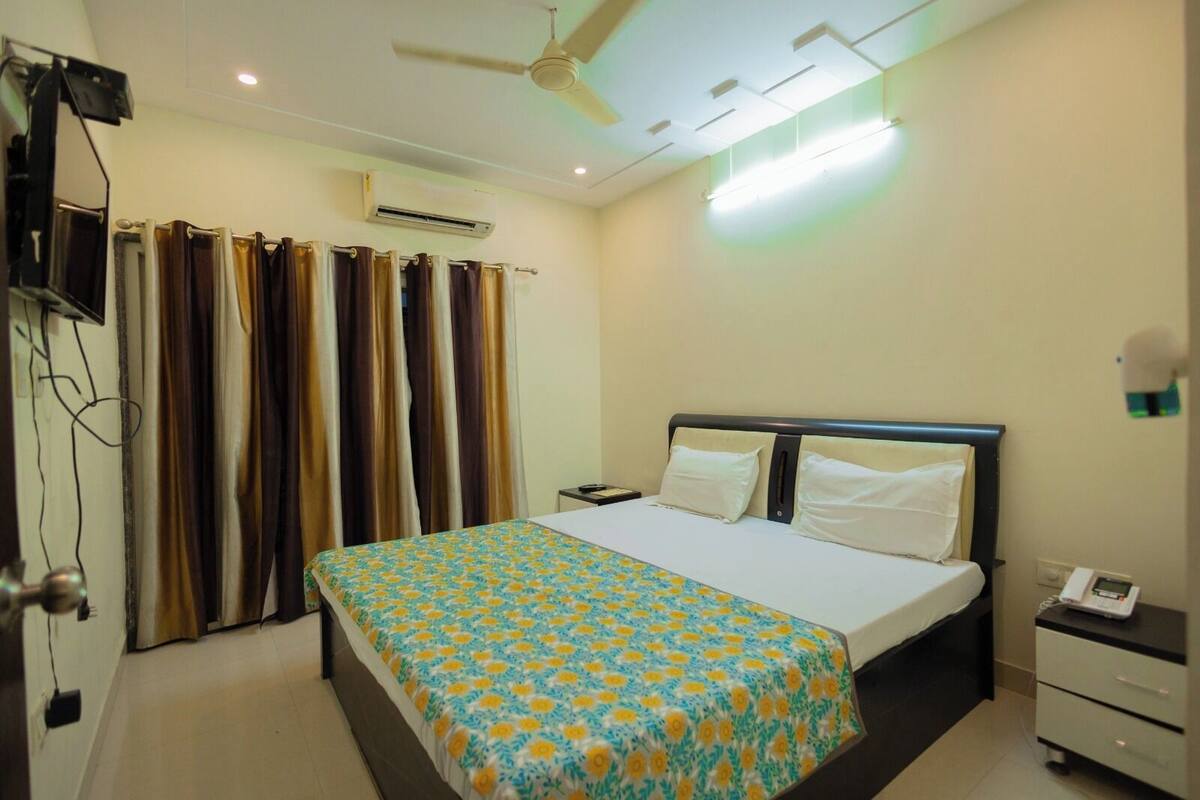 A hotel room with a bed, a bedside table, a nightstand, an air conditioning unit, a ceiling fan, and curtains.