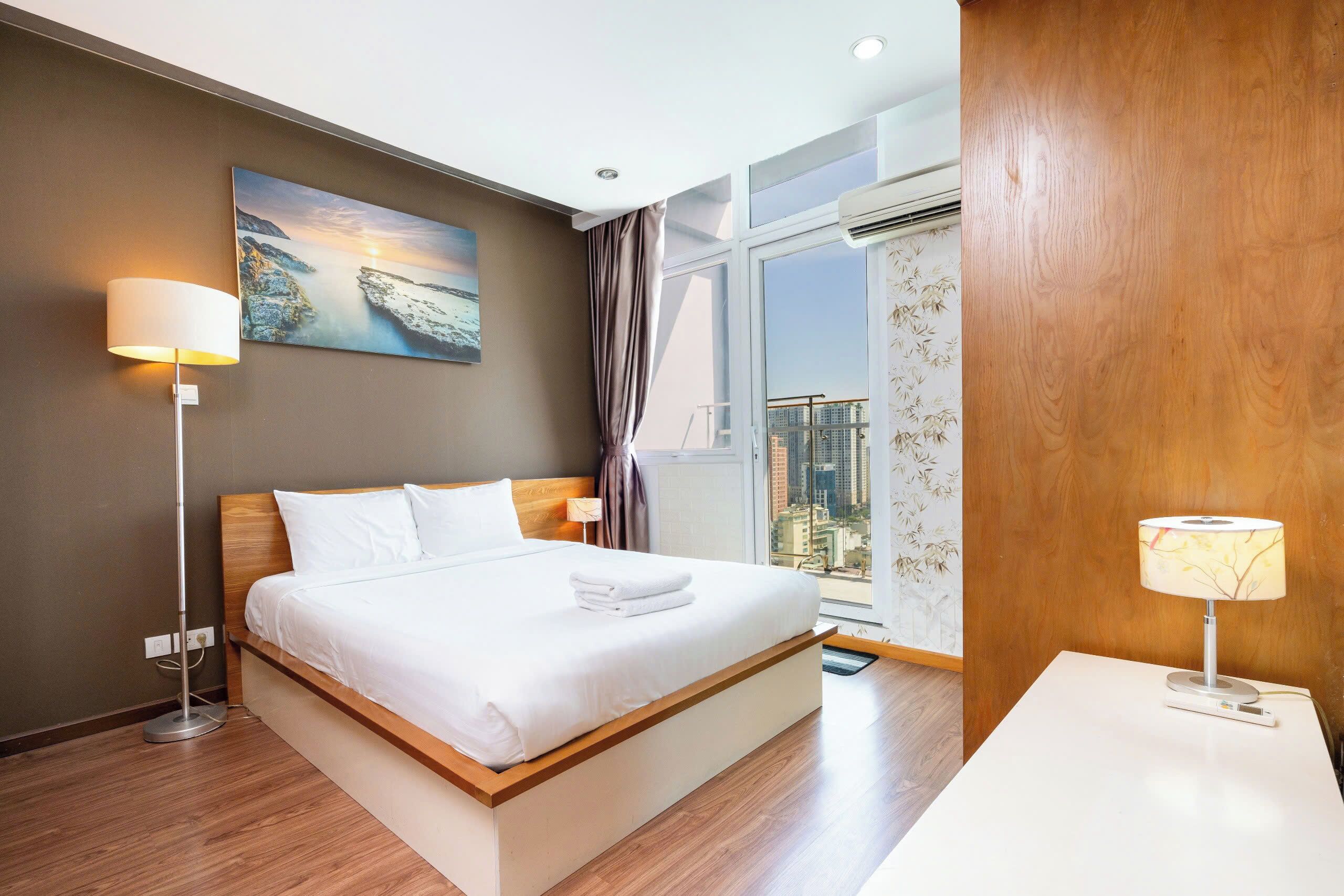 A modern hotel room with a large bed, a nightstand, a floor lamp, a painting of a coastal scene, and a view of a cityscape through a large window.