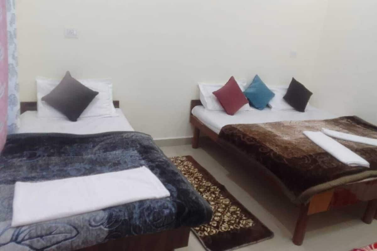 Duas camas de solteiro com almofadas de cores diferentes, uma mesinha e um tapete estampado em um quarto simples, com paredes lisas.