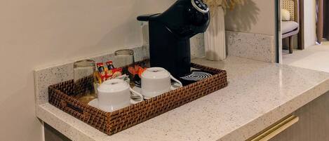 Un comptoir de cuisine avec une machine à café, deux tasses blanches et un panier contenant des bocaux et une bouteille.