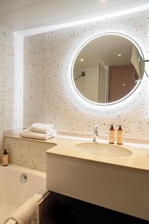 A modern bathroom with a round mirror, a bathtub, and a sink with toiletries. - Mercure Nice Centre Notre Dame Hotel (Nice)