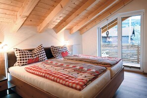 A bedroom with a large bed, checkered pillows, and a view of a wooden deck through a large window. - Cozy and large vacation apartment with all comforts (Sonthofen)