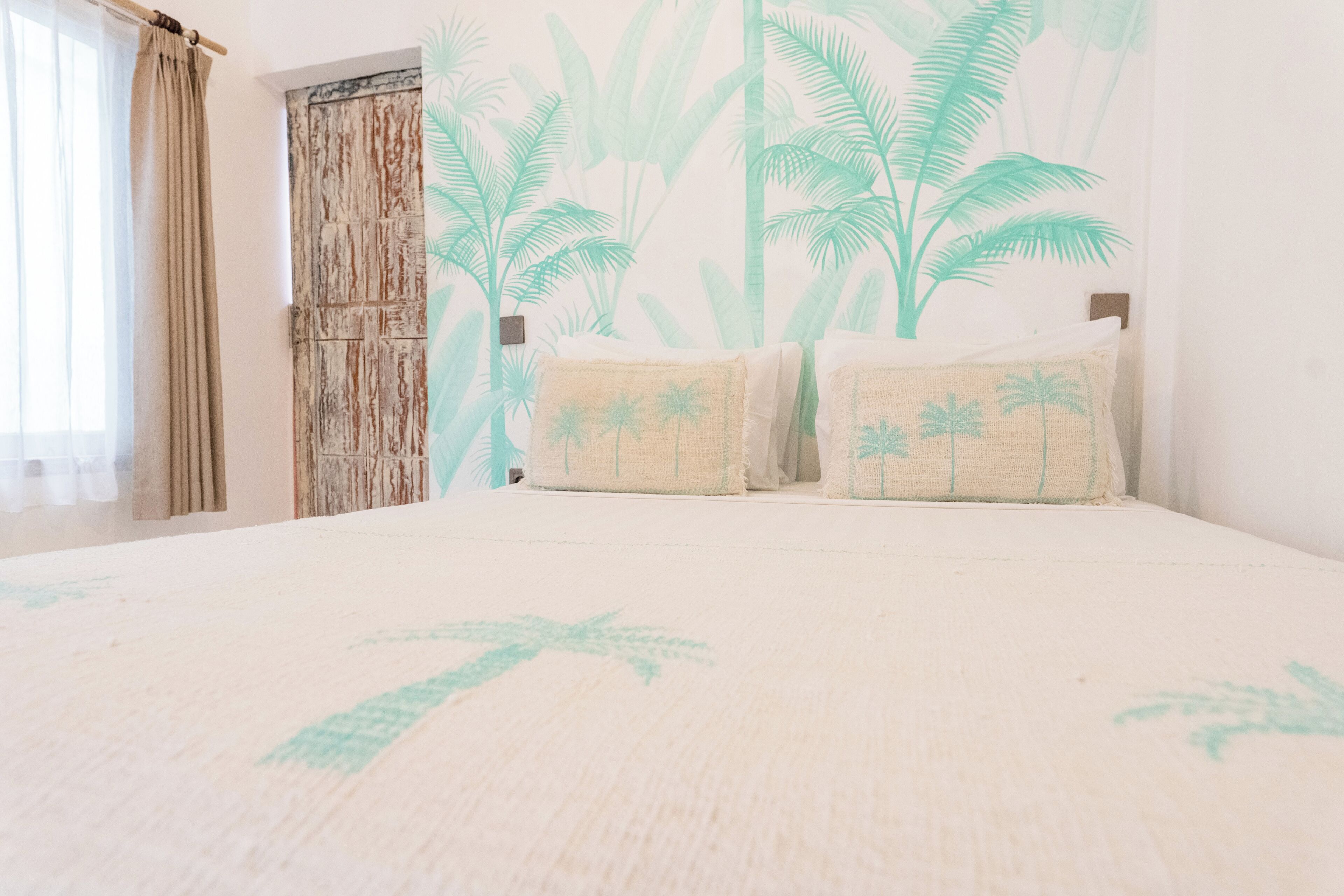 A bedroom with a bed, two pillows with palm tree designs, a wooden door, and a wall with a tropical leaf pattern.