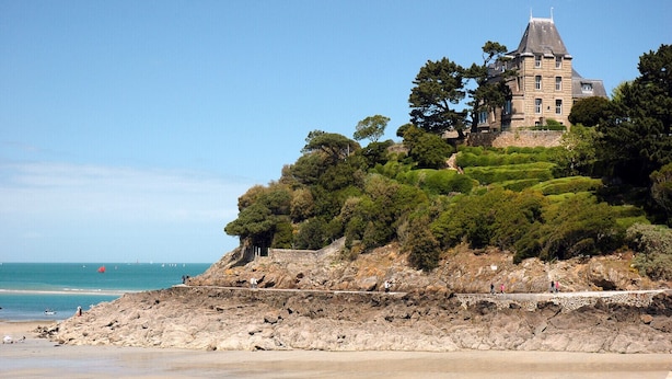 Un bâtiment historique en pierre, doté d’un toit en pente et à plusieurs niveaux, entouré d’une végétation luxuriante et offrant une vue sur une plage.