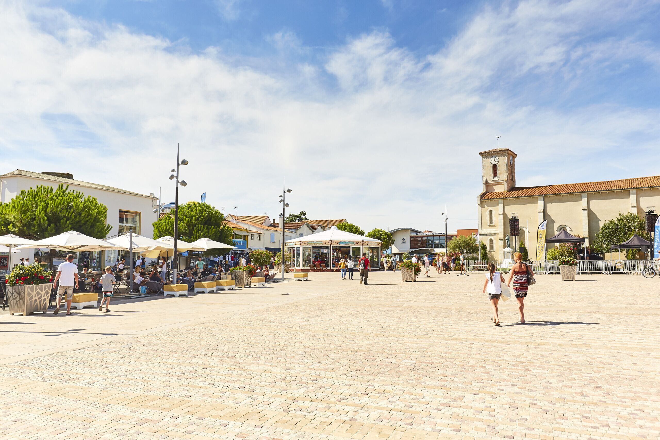 A spacious public square with cobblestone paving, outdoor seating areas, and a prominent clock tower.
