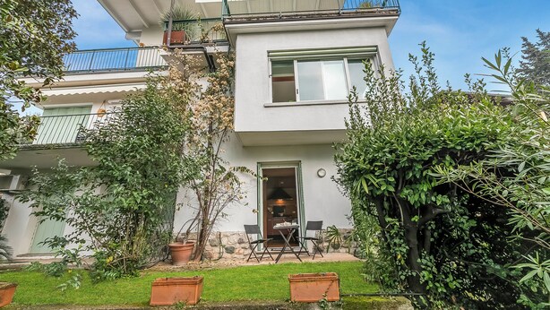 Une maison à deux étages dotée d’un balcon, d’une porte donnant sur un patio avec des chaises et d’un jardin.