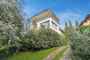 Ein modernes Haus mit einem Glasbalkon, umgeben von üppiger Vegetation und einem Steinweg.