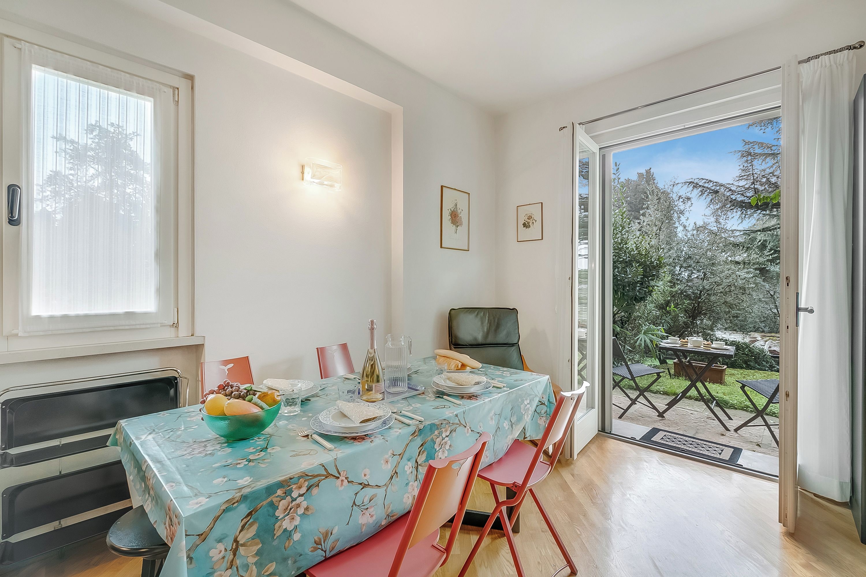 A dining area with a table set for two, a bowl of fruit, and a bottle of wine.