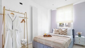 A bedroom with a bed, a bedside table, a nightstand, a window with blinds, and a clothing rack with towels and robes. - Chic 2BR Near Le Louvre & Palais Royal (Paris)
