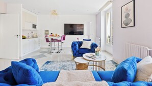A modern living room with a blue sofa, a dining area with a pink chair, and a kitchen with white cabinets. - Chic 2BR Near Le Louvre & Palais Royal (Paris)