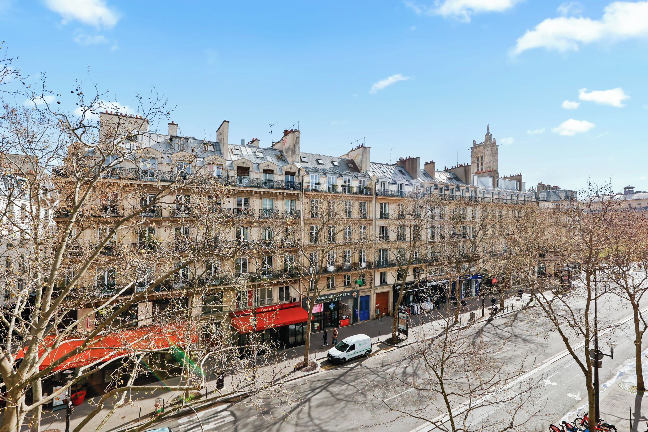A row of historic buildings with multiple stories and balconies, a street with parked cars, and leafless trees.