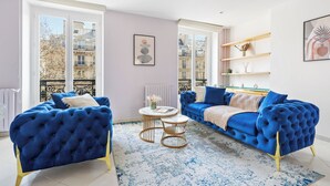 A modern living room with two blue sofas, a round coffee table, and a view of the city through large windows. - Chic 2BR Near Le Louvre & Palais Royal (Paris)