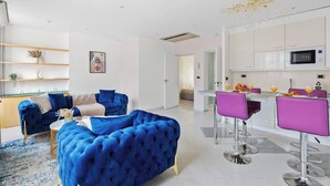 A modern living room with a blue sofa, a coffee table, and a kitchen area with a dining table and bar stools. - Chic 2BR Near Le Louvre & Palais Royal (Paris)