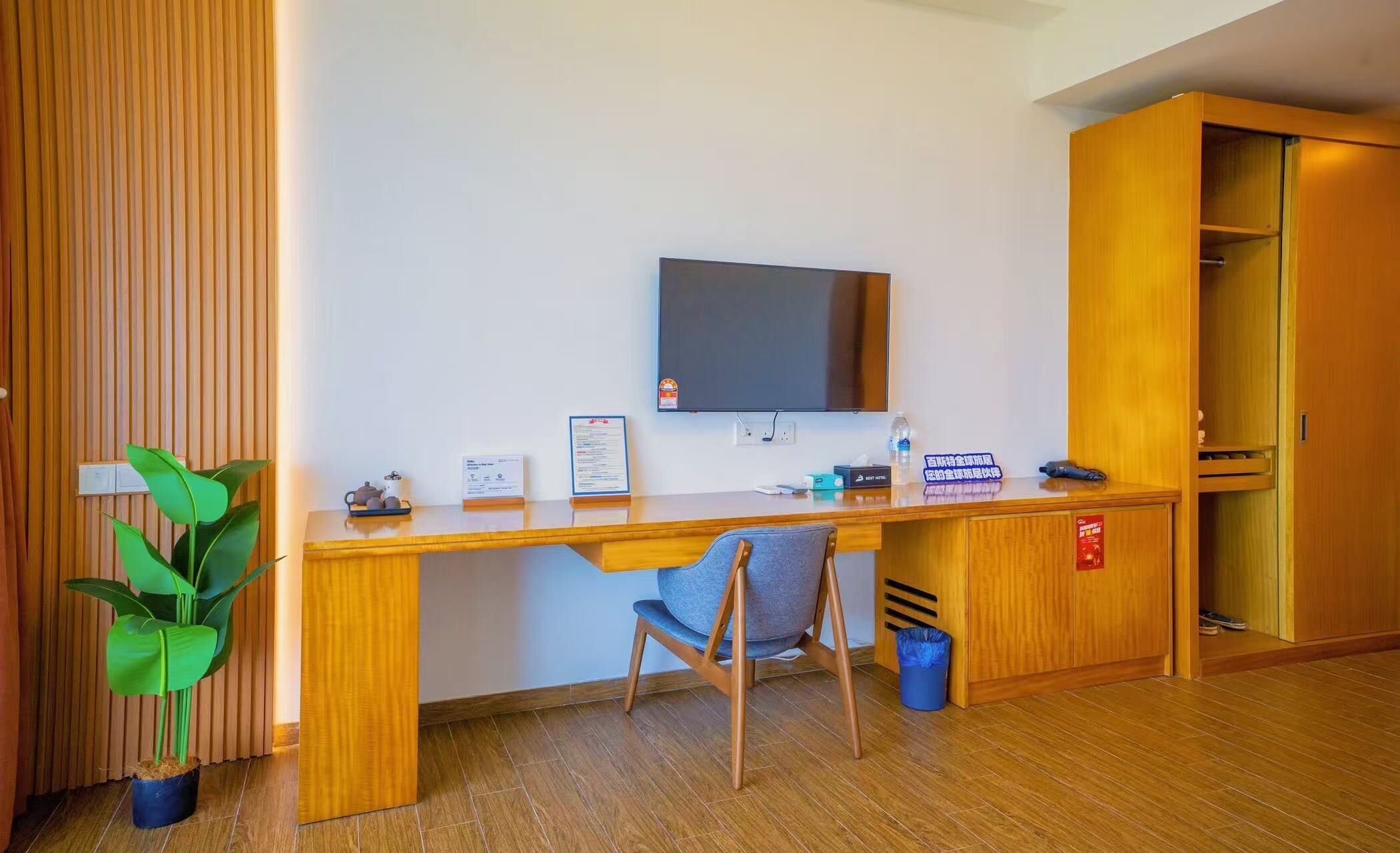 A hotel reception desk with a flat-screen TV, a potted plant, a chair, and a trash bin.