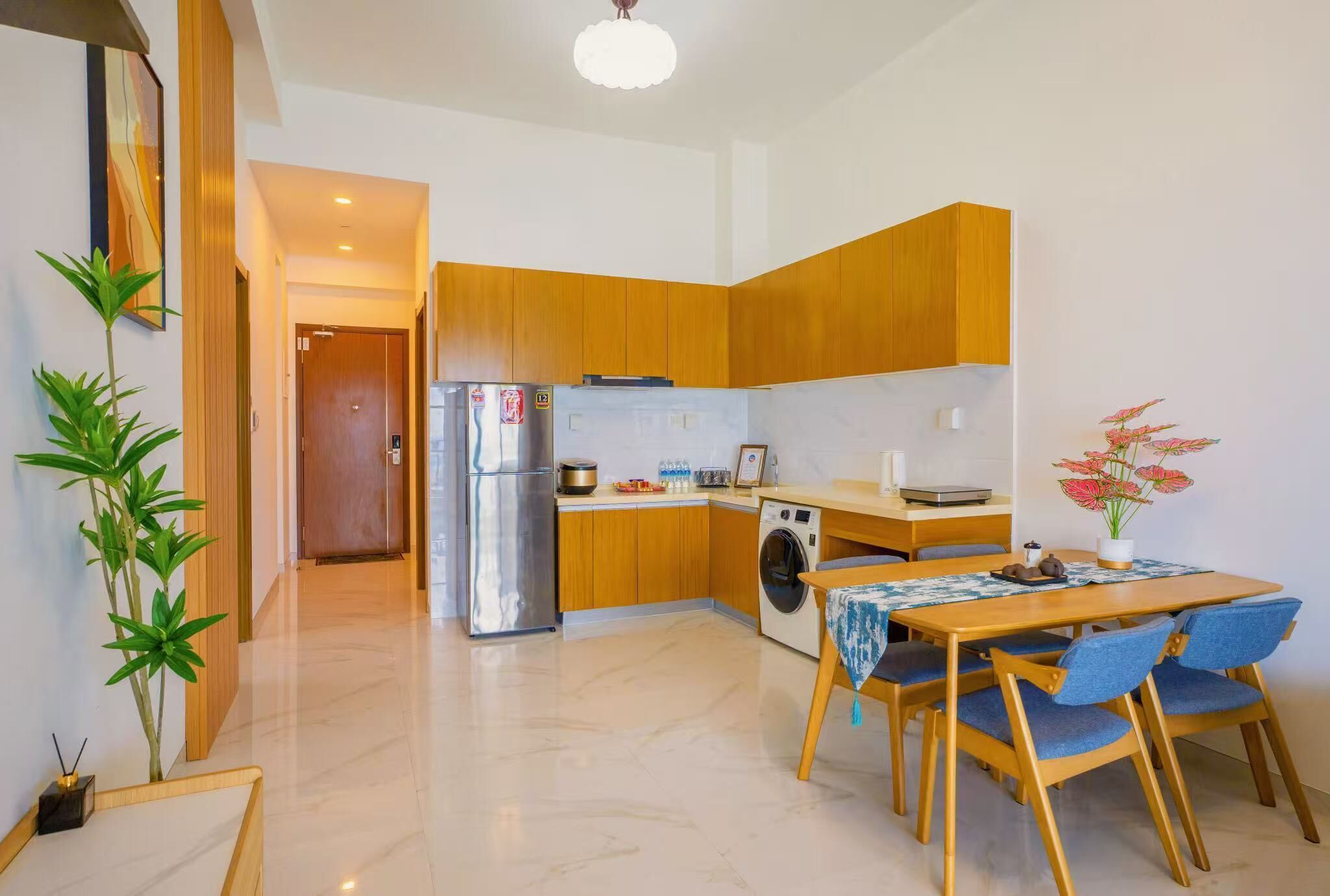 A compact kitchen with wooden cabinets, a refrigerator, a washing machine, and a dining table with chairs.