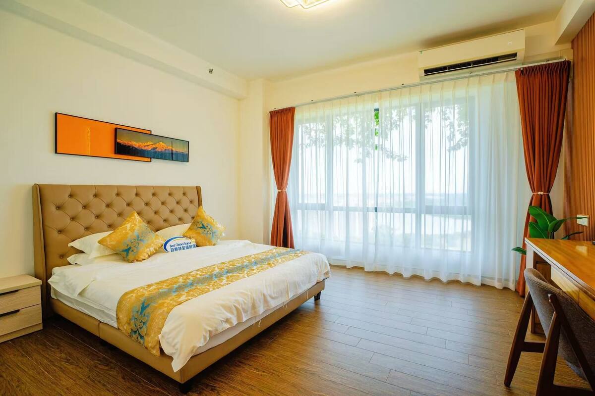 Une chambre avec un grand lit, une table de chevet, un bureau, une chaise, une fenêtre avec des rideaux et un tableau au mur.