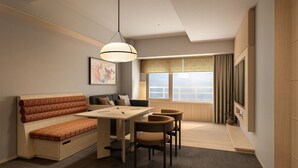 Een moderne woonkamer met een bank, eettafel, stoelen, een hanglamp, een groot raam met jaloezieën en een televisie.
