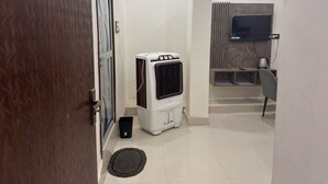 A hotel room with a white air conditioner, a black trash bin, a black doormat, a tiled floor, a wooden door, and a wall-mounted TV. - Shri Awadh Hotel (Sitamarhi)