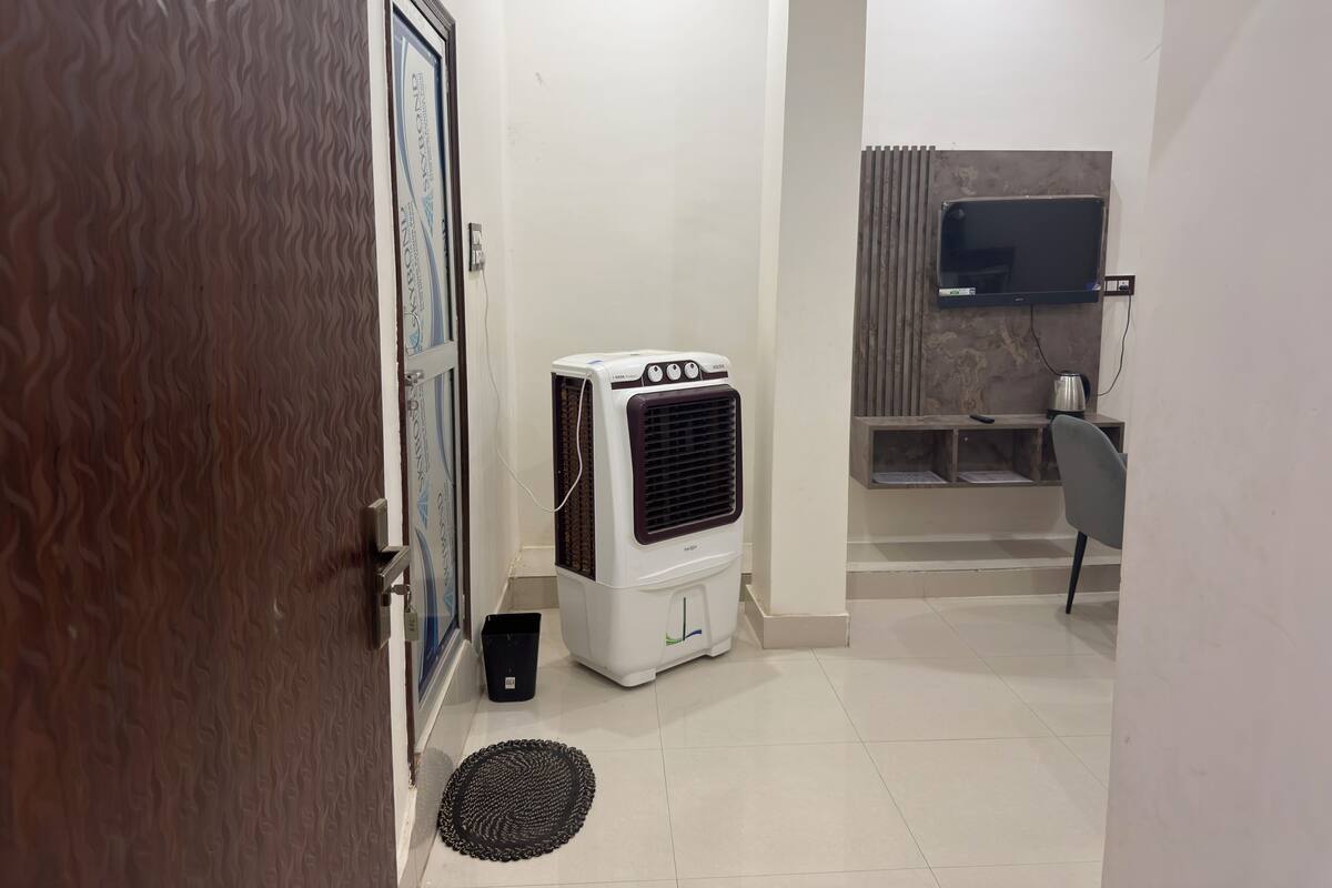 Habitación de hotel con aire acondicionado blanco, un cesto de basura negro, felpudo negro, suelo de baldosas, puerta de madera y televisor montado en la pared.