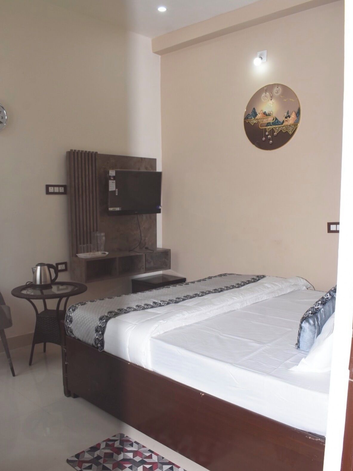 Un dormitorio con cama, televisor montado en la pared, una mesita con tetera y un adorno en la pared.