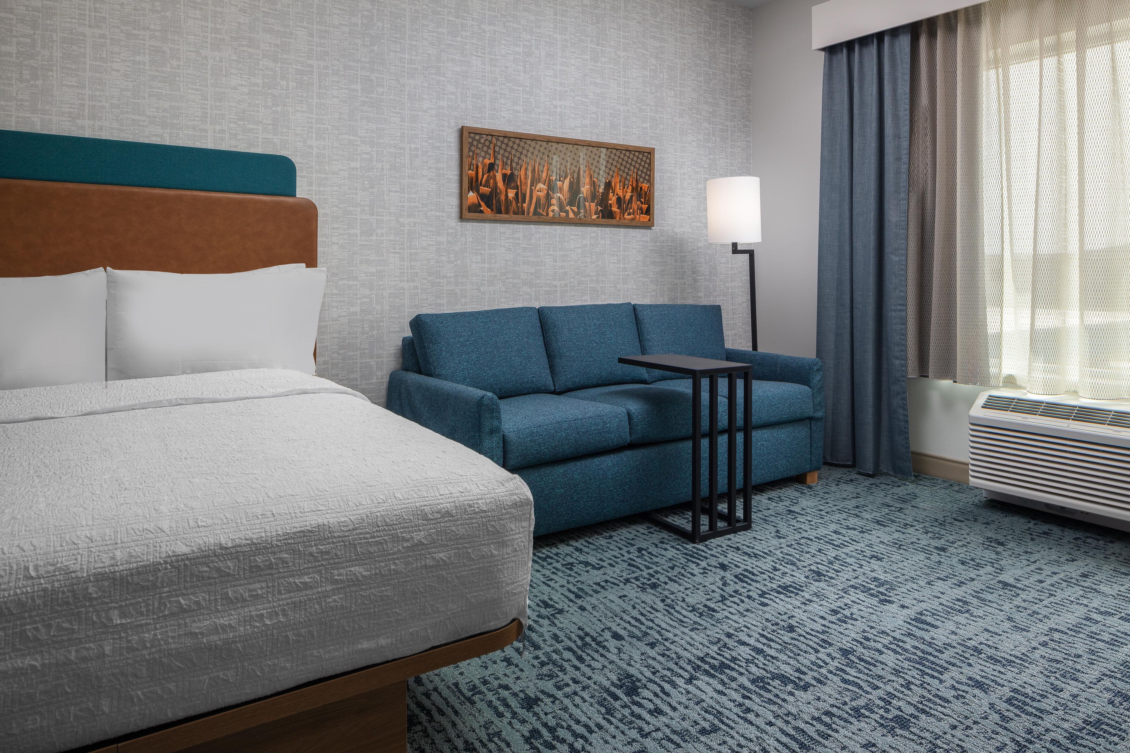 A hotel room with a bed, a blue sofa, a small table, a lamp, and a large window with curtains.
