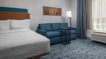 A hotel room with a bed, a blue sofa, a small table, a lamp, and a large window with curtains.