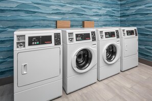 A row of white front-loading washers with digital displays, set against a blue wavy wall. - Hampton Inn by Hilton Grand Island Medical Center (Grand Island)