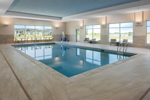 A large indoor swimming pool with clear views of the outside, surrounded by tiled flooring and equipped with sun loungers and chairs.