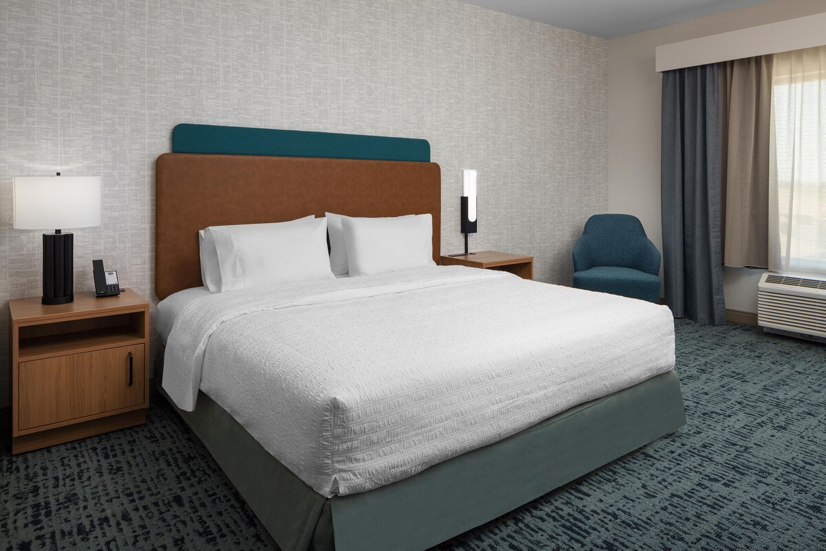 A hotel room with a large bed, a nightstand with a lamp, a chair, and a window with curtains.
