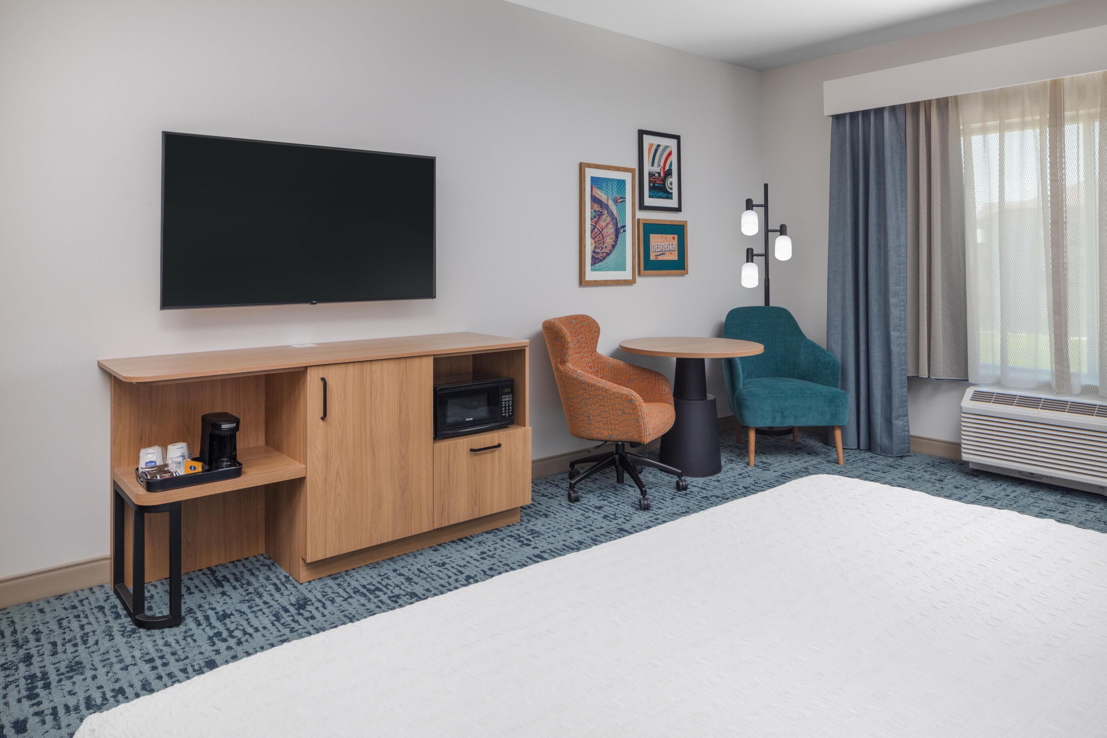 A hotel room with a bed, a TV, a desk with a microwave, a coffee maker, two chairs, and a heater.
