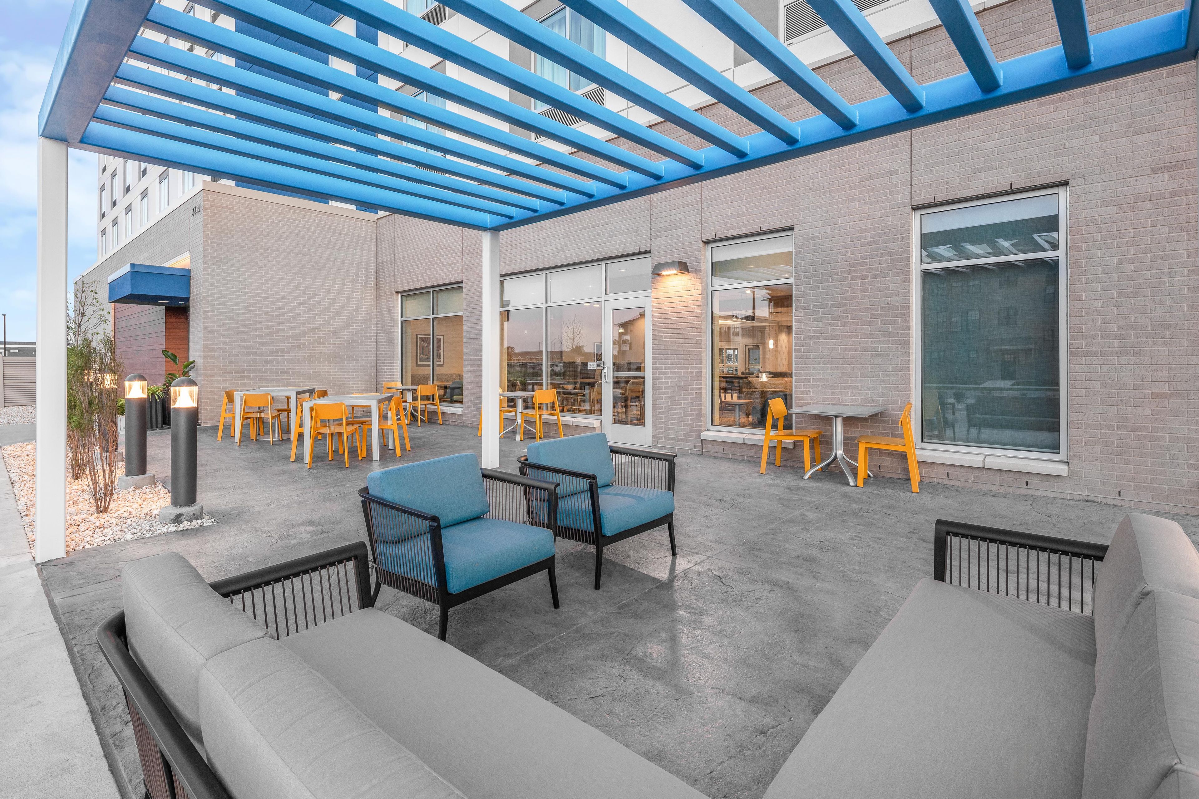 A covered outdoor seating area with chairs and tables, a brick building with large windows, and a blue pergola structure.