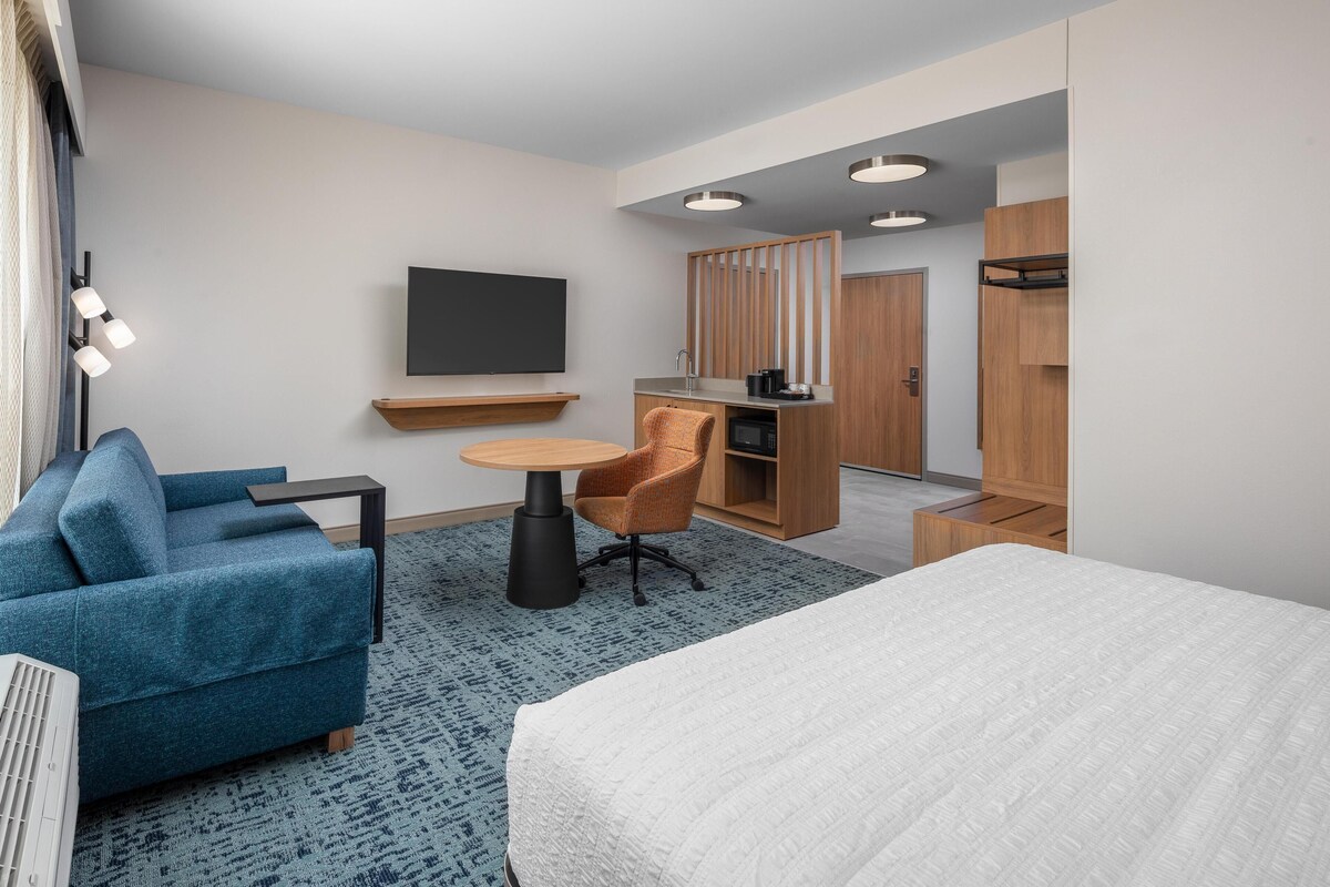 A modern hotel room with a bed, a blue sofa, a small round table, a TV, a kitchenette, and a wardrobe.