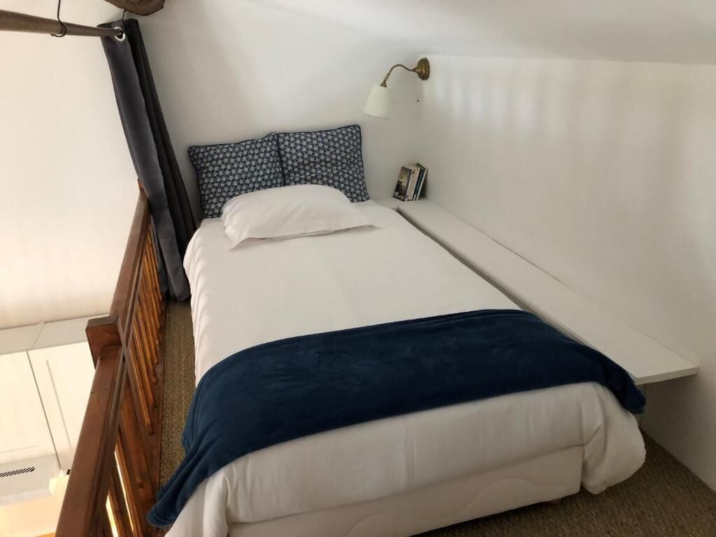 Ein Einzelbett mit weißer Matratze, einer blauen Decke und einem gemusterten Kopfkissen. Ein Nachttisch mit Lampe ist sichtbar.