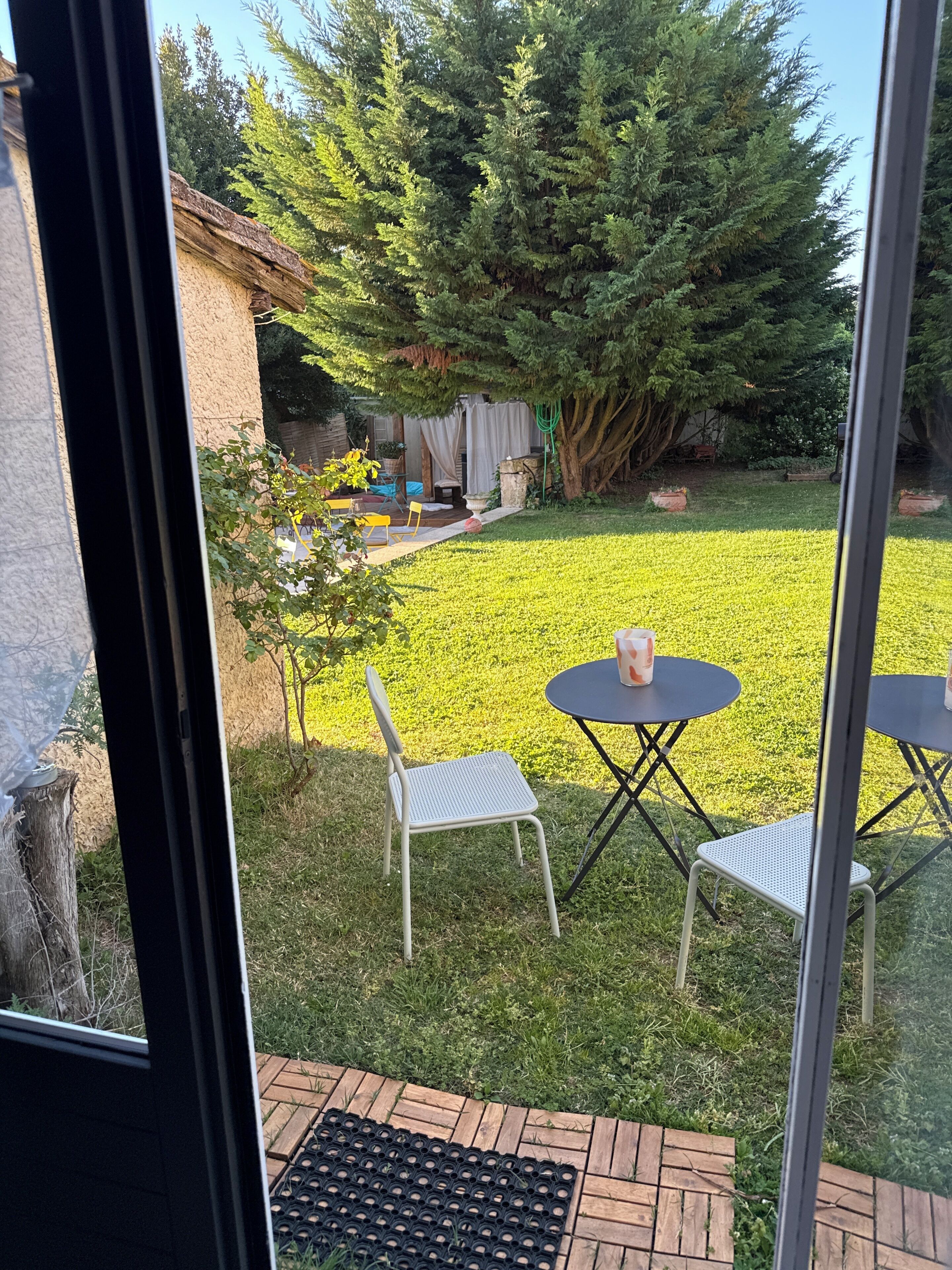 Une terrasse avec deux tables rondes et deux chaises, un petit jardin avec de l’herbe et un arbre, et une vue sur une maison avec une cheminée.