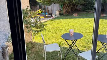 Une terrasse avec deux tables rondes et deux chaises, un petit jardin avec de l’herbe et un arbre, et une vue sur une maison avec une cheminée.