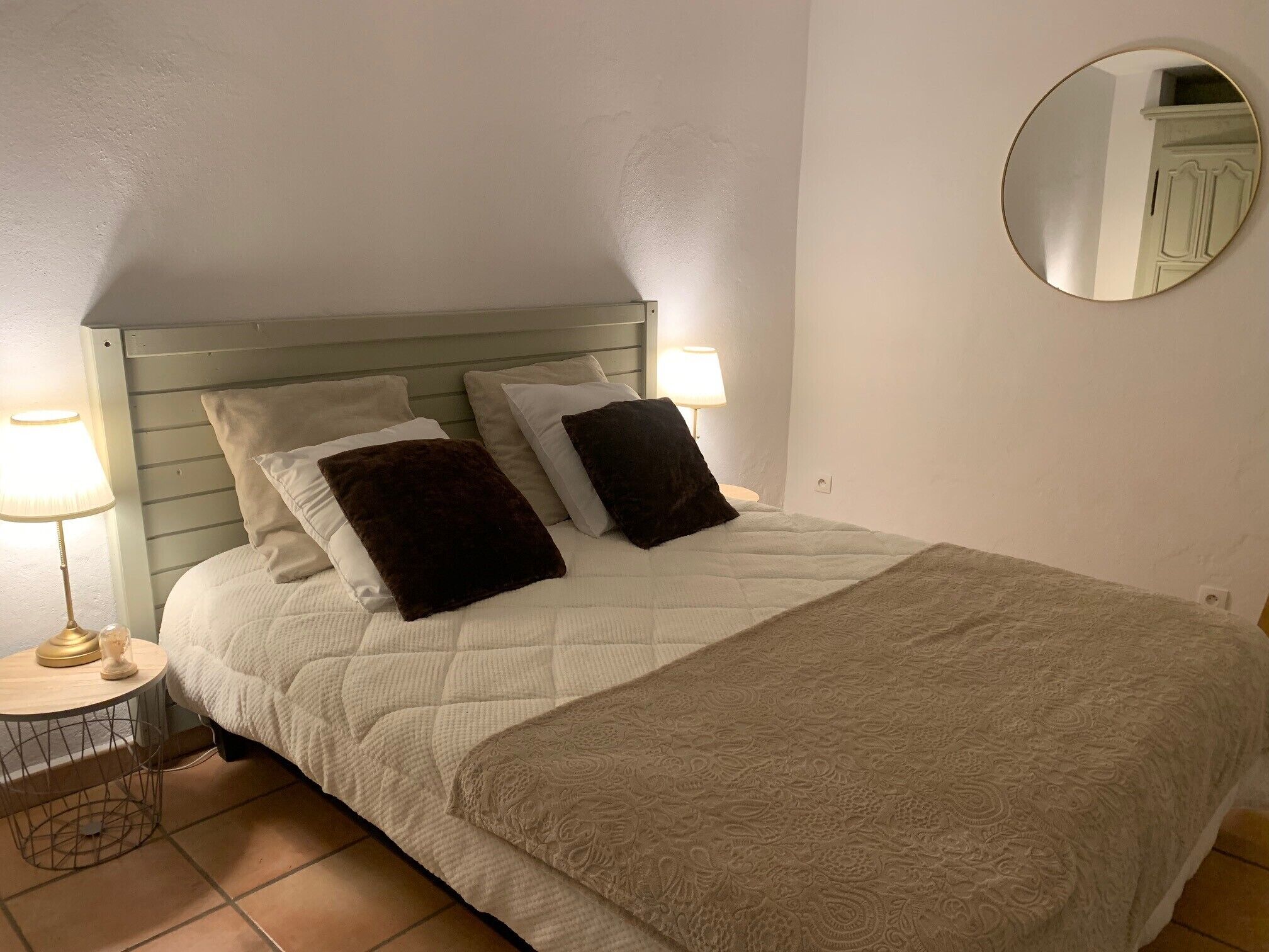 A neatly made bed with a quilted white duvet and a patterned throw blanket. There are two bedside lamps on a small round table beside the bed.