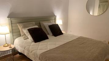 A neatly made bed with a quilted white duvet and a patterned throw blanket. There are two bedside lamps on a small round table beside the bed.