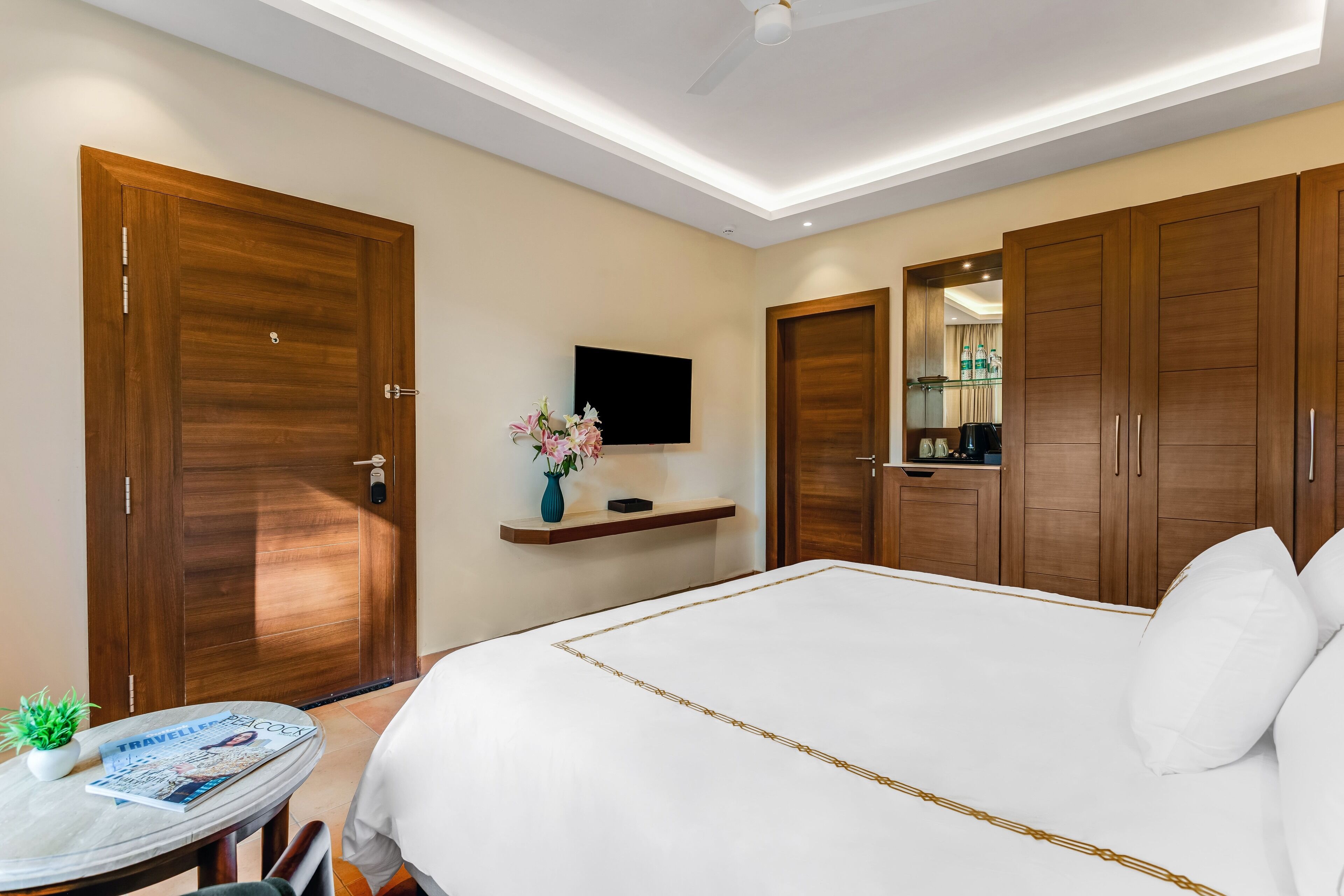 Habitación de hotel con una cama grande, armarios de madera, televisor montado y una mesita con planta y revista.