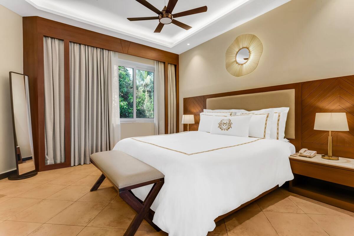 A hotel room with a large bed, a bench, a ceiling fan, a bedside table with a lamp, and a view of greenery outside.